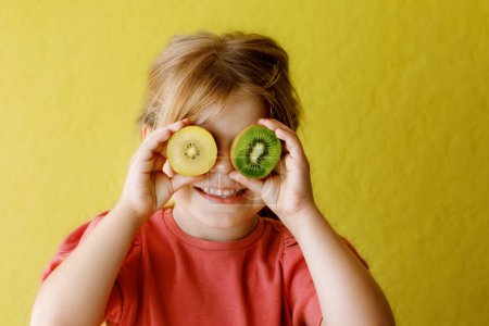 Téléchargez les photos : Happy little child girl with yellow and green kiwi fruits on yellow background. Preschool girl smiling. Healthy fruits for children. - en image libre de droit