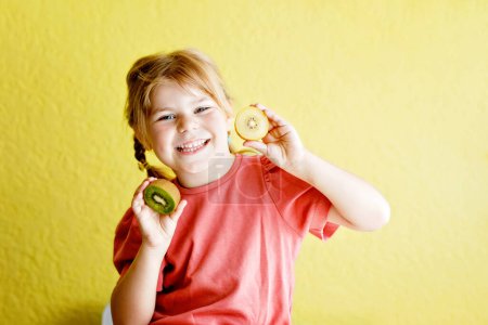 Foto de Happy little child girl with yellow and green kiwi fruits on yellow background. Preschool girl smiling. Healthy fruits for children. - Imagen libre de derechos