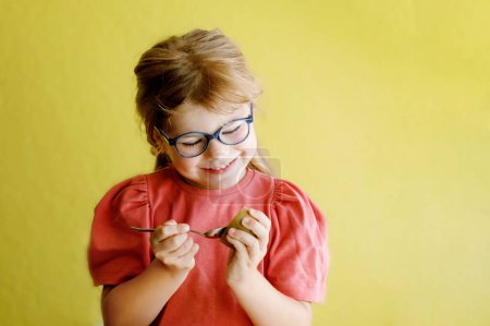 Foto de Niña feliz con gafas comiendo kiwi con cuchara sobre fondo amarillo. Una niña en edad preescolar sonriendo. Frutas saludables para los niños. - Imagen libre de derechos