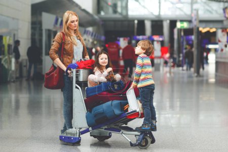 Photo for Two little kids, boy and girl, siblings and mother at the airport. Children, family traveling, going on vacation by plane and waiting on trolley with suitcases pushing by woman at terminal for flight - Royalty Free Image