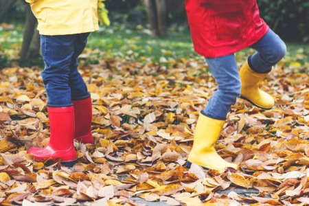 Foto de Dos niños pequeños jugando con botas de goma rojas y amarillas en el parque de otoño con coloridos abrigos y ropa de lluvia. Primer plano de las piernas de los niños en zapatos bailando y caminando a través de las hojas otoñales de otoño y el follaje - Imagen libre de derechos