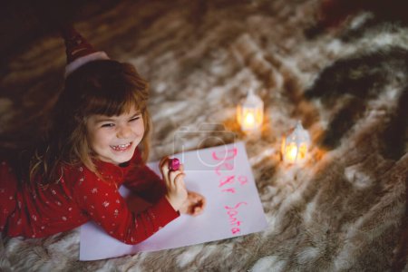 Téléchargez les photos : Petite fille mignonne préscolaire en pyjama sous l'arbre de Noël, écrivant lettre de liste de souhaits au Père Noël à la maison, à l'intérieur. Fête chrétienne traditionnelle. Bébé heureux enfant en attente de cadeaux sur Noël - en image libre de droit