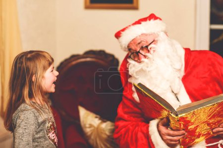 Photo for Little toddler girl talking to Santa Claus called Nikolaus or Weihnachtsmann in German. Happy smiling kid waiting for gifts. Santa with golden book. Xmas, childhood, holiday. - Royalty Free Image