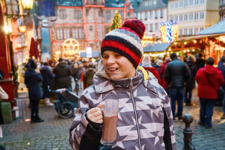 Téléchargez les photos : Petit garçon mignon qui boit du punch ou du chocolat chaud pour enfants sur le marché de Noël allemand. Enfant heureux sur le marché familial traditionnel en Allemagne, Rire garçon en vêtements d'hiver colorés. - en image libre de droit
