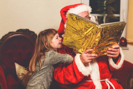 Foto de Niña pequeña hablando con Santa Claus llamada Nikolaus o Weihnachtsmann en alemán. Feliz niño sonriente esperando regalos. Santa con libro de oro. Navidad, infancia, vacaciones. - Imagen libre de derechos