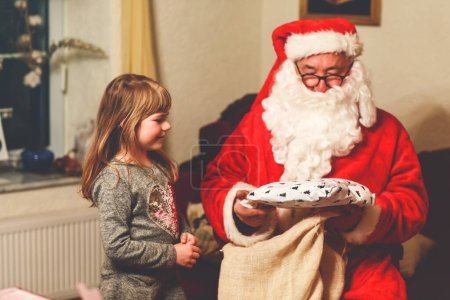 Photo for Little toddler girl talking to Santa Claus called Nikolaus or Weihnachtsmann in German. Happy smiling kid waiting for gifts. Santa with golden book. Xmas, childhood, holiday. - Royalty Free Image