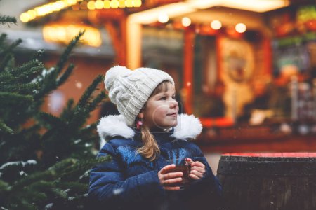 Téléchargez les photos : Petite fille préscolaire mignonne qui boit du punch chaud pour enfants ou du chocolat sur le marché de Noël allemand. Enfant heureux sur le marché familial traditionnel en Allemagne, Rire garçon en vêtements d'hiver colorés. - en image libre de droit