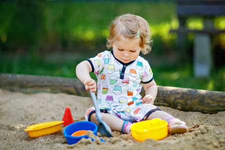 Foto de Linda niña jugando en la arena en el patio al aire libre. Hermoso bebé en ropa de verano que se divierte en el soleado día cálido de verano. Actividad al aire libre para niños pequeños - Imagen libre de derechos