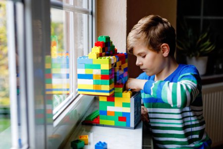 Téléchargez les photos : Petit garçon jouant avec beaucoup de blocs en plastique coloré. Adorable écolier s'amuser avec la construction et la création bâtiment par fenêtre. Technique créative de loisirs et robotique pendant le temps de couronne. - en image libre de droit