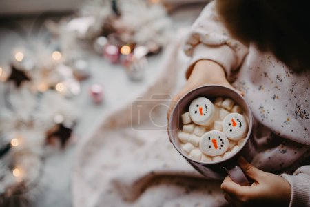 Téléchargez les photos : Petite fille méconnaissable tenant une tasse de chocolat chaud avec des guimauves comme bonhomme de neige. Enfant assis près de Noël décoré fenêtre avec des lumières - en image libre de droit
