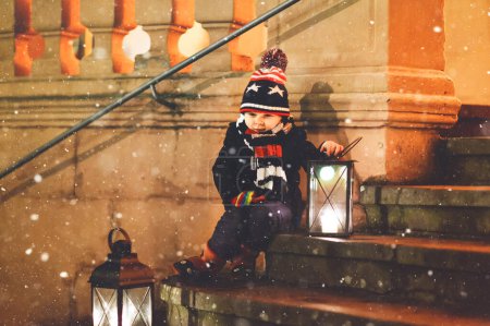 Foto de Niño lindo con una linterna de luz en las escaleras cerca de la iglesia. Niño feliz en el mercado de Navidad en Alemania. Niño esperando a los padres en el frío día de invierno. - Imagen libre de derechos