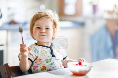 Niña adorable comer de cuchara fresca saludable frambuesas alimentos, niño, alimentación y concepto de desarrollo. Lindo niño, hija con cuchara sentada en silla alta y aprender a comer por sí mismo
