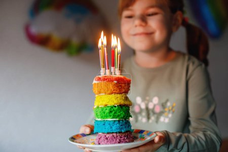 Foto de Happy little preschool girl celebrating birthday. Cute smiling child with homemade rainbow cake, indoor. Happy healthy toddler blowing six candles on cake. Selective focus on cake. - Imagen libre de derechos