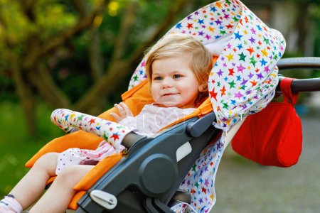 Foto de Retrato de una niña pequeña y sonriente sentada en un cochecito o cochecito de bebé y dando un paseo. Feliz bebé lindo que se divierte al aire libre. Hija sana - Imagen libre de derechos