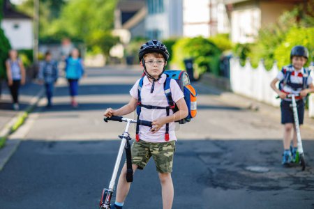 Photo for Two school kid boys in safety helmet riding with scooter in the city with backpack on sunny day. Happy children in colorful clothes biking on way to school. Safe way for kids outdoors to school. - Royalty Free Image