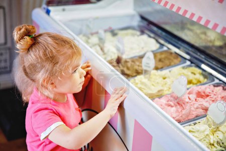 Photo for Cute little toddler girl choosing and buying ice cream in a cafe. Happy baby child looking at different sorts of icecream. Sweet home made dessert. - Royalty Free Image