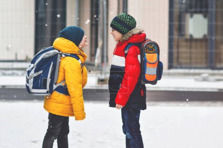 Foto de Dos niños pequeños de clase primaria caminando a la escuela durante las nevadas. Niños felices divirtiéndose y jugando con la primera nieve. Hermanos y mejores amigos con mochila en ropa de invierno colorida - Imagen libre de derechos