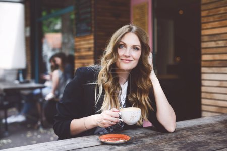Téléchargez les photos : Jeune femme d'affaires buvant du cappuccino et une tasse de café. Heureuse femme seule dans un café ou un restaurant en plein air le jour d'été ensoleillé, pause pour le déjeuner entre les réunions - en image libre de droit