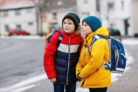 Foto de Dos niños pequeños de clase primaria caminando a la escuela durante las nevadas. Niños felices divirtiéndose y jugando con la primera nieve. Hermanos y mejores amigos con mochila en ropa de invierno colorida - Imagen libre de derechos