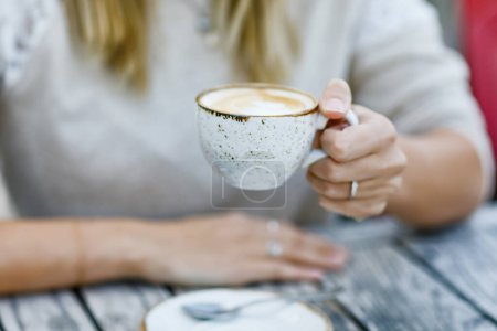 Foto de Primer plano de las manos de la mujer que bebe capuchino y taza de café mientras espera a amigo y desayuno. Feliz mujer sola en la cafetería al aire libre o restaurante en el soleado día de verano - Imagen libre de derechos