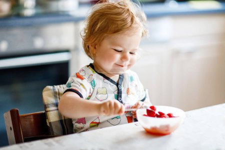 Photo for Adorable baby girl eating from spoon fresh healthy raspberries food, child, feeding and development concept. Cute toddler, daughter with spoon sitting in highchair and learning to eat by itself - Royalty Free Image