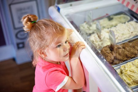 Téléchargez les photos : Petite fille mignonne tout-petit choisir et acheter de la crème glacée dans un café. Bébé enfant heureux regardant différentes sortes de glaçons. Sucré dessert fait maison
. - en image libre de droit