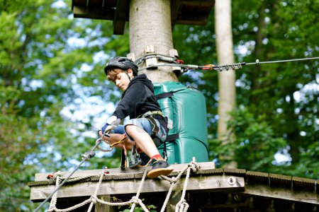 Photo for School boy in forest adventure park. Acitve child, kid in helmet climbs on high rope trail. Agility skills and climbing outdoor amusement center for children. Outdoors activity for kid and families - Royalty Free Image