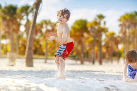 Téléchargez les photos : Adorable petit garçon actif qui s'amuse sur la plage de Miami, Key Biscayne. Joyeux enfant mignon relaxant, jouant avec le sable et profitant d'une journée chaude ensoleillée près des paumes et de l'océan - en image libre de droit