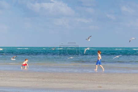 Foto de Dos niños pequeños divirtiéndose en la playa tropical, felices mejores amigos jugando, concepto de amistad. Hermanos hermanos corriendo en el agua y la captura de aves gaviotas. Vacaciones familiares en el océano - Imagen libre de derechos