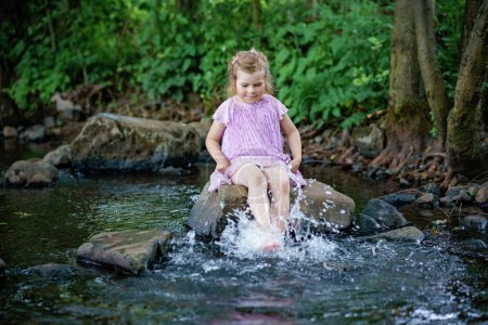 Foto de Linda niña pequeña divirtiéndose junto a un río en un día de verano cálido y soleado. Feliz niño preescolar emocionado salpicando con agua en arroyo forestal - Imagen libre de derechos