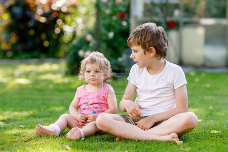 Foto de Happy little kid boy with cute little baby girl, cute sister. Siblings. Brother and baby playing together in summer garden with flowers. Family and love. - Imagen libre de derechos
