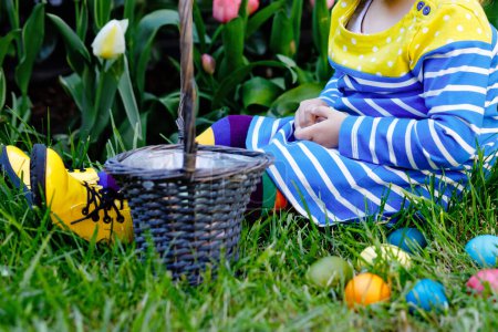 Photo for Cute little toddler girl with bunny ears having fun with traditional Easter eggs hunt on warm sunny day, outdoors. Happy child celebrating family christian holiday with basket with colored egg. - Royalty Free Image