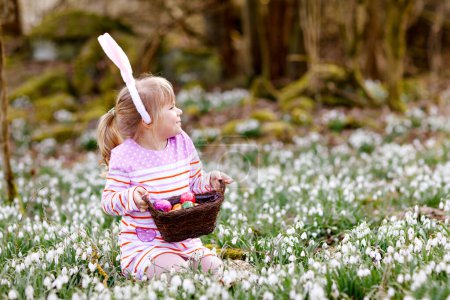 Foto de Niña con orejas de conejo de Pascua haciendo caza de huevos en el bosque de primavera en un día soleado, al aire libre. Lindo niño feliz con un montón de flores de nieve y huevos de colores. Primavera, concepto de vacaciones cristianas - Imagen libre de derechos