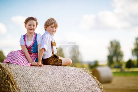 Téléchargez les photos : Deux enfants en costumes bavarois traditionnels dans le champ de blé. Des enfants allemands assis sur une balle de foin pendant l'Oktoberfest. Garçon et fille jouent aux balles de foin pendant la récolte estivale en Allemagne. Meilleurs amis - en image libre de droit