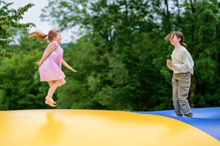 Téléchargez les photos : Petite fille d'âge préscolaire et sœur d'école sautant sur le trampoline. Heureux enfants drôles, frères et sœurs amoureux s'amuser avec l'activité de plein air en été. Trampoline aux couleurs du drapeau ukrainien. - en image libre de droit