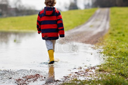 Foto de Niño pequeño que usa botas de lluvia amarillas y camina y salta al charco en un cálido y soleado día de primavera. Niño feliz en ropa de lluvia casual de moda colorida divertirse y jugar al aire libre - Imagen libre de derechos