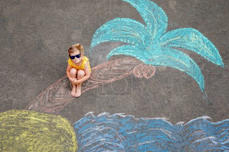 Téléchargez les photos : Happy little preschool girl in swimsuit on inflatable ring with sea, sand, palm painted with colorful chalks on asphalt. Cute child with having fun with chalk picture. Summer, vacations, summertime. - en image libre de droit