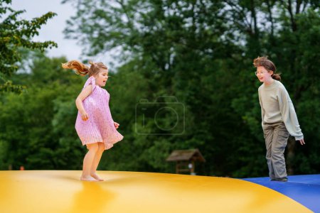 Photo for Little preschool girl and school sister jumping on trampoline. Happy funny children, siblings in love having fun with outdoor activity in summer. Trampolin in ukrainian flagg colors. - Royalty Free Image