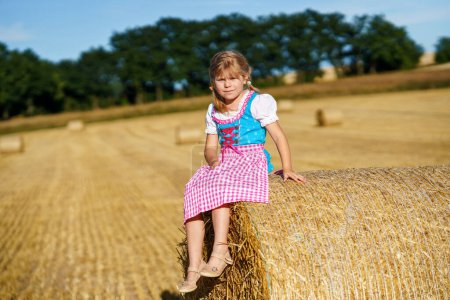 Foto de Cute little kid girl in traditional Bavarian costume in wheat field. Happy child with hay bale during Oktoberfest in Munich. Preschool girl play at hay bales during summer harvest time in Germany - Imagen libre de derechos