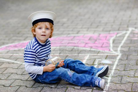 Téléchargez les photos : Petit garçon comme pirate sur le bateau ou voilier peinture d'image avec des craies colorées sur asphalte. Loisirs créatifs pour les enfants à l'extérieur en été. Enfant avec chapeau de capitaine et jumelles - en image libre de droit