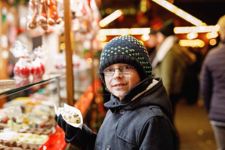 Téléchargez les photos : Petit garçon enfant, mignon enfant mangeant des bananes recouvertes de chocolat, de marshmellows et de saupoudres colorées près d'un stand sucré avec du pain d'épice et des noix. Joyeux garçon sur le marché de Noël en Allemagne - en image libre de droit