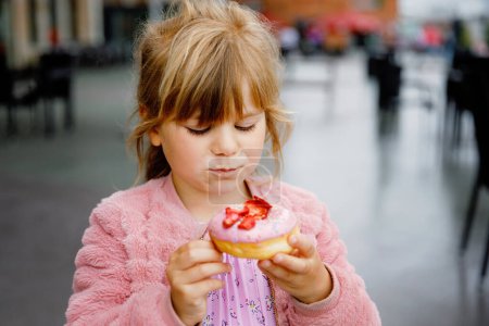 Photo for Happy little preschool girl eating sweet donut indoor. Blond child in the city. Cute kid and unhealthy sugared food doughnut - Royalty Free Image