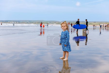 Téléchargez les photos : Petite fille mignonne tout-petit à la plage de surf Ballybunion, s'amusant à jouer sur la côte ouest de l'Irlande. Enfant heureux profitant de l'été irlandais et de la journée ensoleillée en famille - en image libre de droit