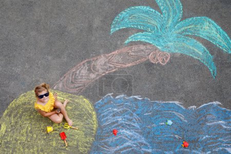 Téléchargez les photos : Happy little preschool girl in swimsuit with sea, sand, palm painted with colorful chalks on asphalt. Cute child with having fun with chalk picture. Summer, vacations, summertime. - en image libre de droit