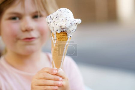 Foto de Pequeña niña preescolar adorable comiendo helado en cono de gofre en el día soleado de verano. Feliz niño pequeño come postre helado. Comida dulce en días calurosos de verano. - Imagen libre de derechos