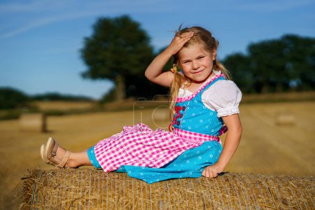 Photo for Cute little kid girl in traditional Bavarian costume in wheat field. Happy child with hay bale during Oktoberfest in Munich. Preschool girl play at hay bales during summer harvest time in Germany - Royalty Free Image