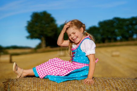 Téléchargez les photos : Cute little kid girl in traditional Bavarian costume in wheat field. Happy child with hay bale during Oktoberfest in Munich. Preschool girl play at hay bales during summer harvest time in Germany - en image libre de droit