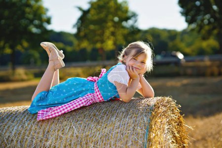 Foto de Cute little kid girl in traditional Bavarian costume in wheat field. Happy child with hay bale during Oktoberfest in Munich. Preschool girl play at hay bales during summer harvest time in Germany - Imagen libre de derechos