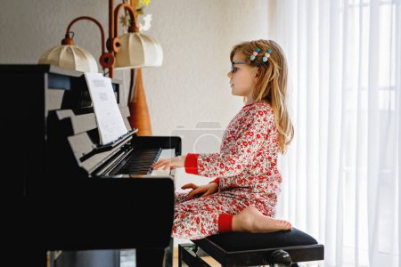 Photo for Little happy girl playing piano in living room. Cute preschool child with eye glasses having fun with learning to play music instrument - Royalty Free Image