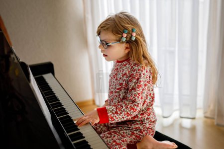 Téléchargez les photos : Little happy girl playing piano in living room. Cute preschool child with eye glasses having fun with learning to play music instrument - en image libre de droit